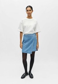 Femme debout devant un fond uni portant un t-shirt blanc oversized, une jupe portefeuille en denim bleu, des collants noirs et des mocassins noirs.