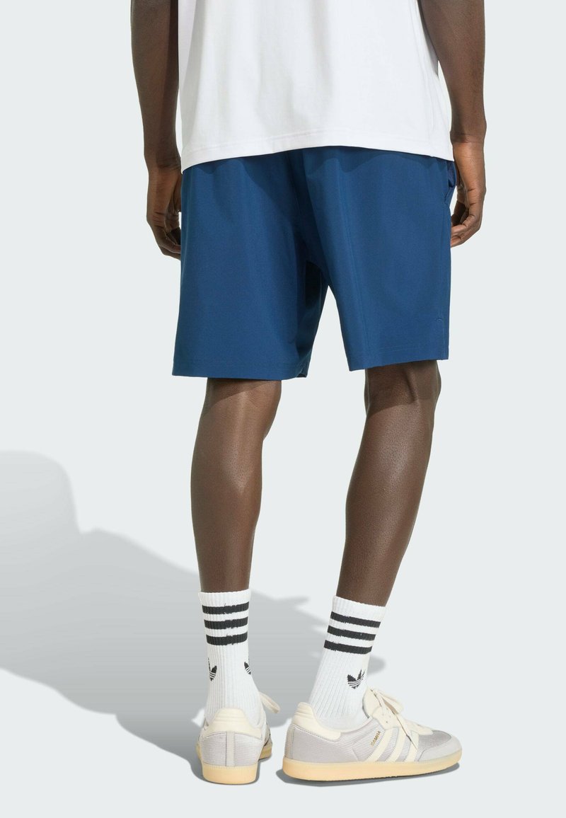 Mann trägt marineblaue Shorts, weiße Adidas-Socken mit drei schwarzen Streifen und weiße Adidas-Sneaker, steht mit leicht geöffneten Beinen.