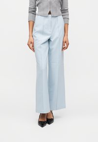 Γυναίκα που φοράει ψηλόμεσα ανοιχτό μπλε παντελόνια με φαρδύ πόδι, γκρι κοντό πουλόβερ και μαύρες μυτερές γόβες, στέκεται μπροστά σε λευκό φόντο.