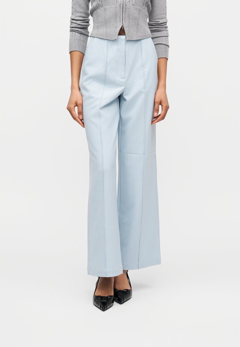 Γυναίκα που φοράει ψηλόμεσα ανοιχτό μπλε παντελόνια με φαρδύ πόδι, γκρι κοντό πουλόβερ και μαύρες μυτερές γόβες, στέκεται μπροστά σε λευκό φόντο.