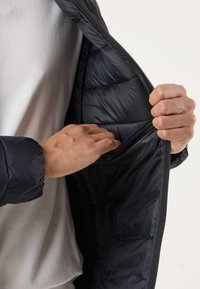 Personne portant une chemise blanche qui déboutonne une veste matelassée noire et atteint la poche intérieure du côté gauche.