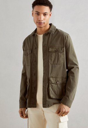 Chaqueta de botones en verde oliva con cuatro bolsillos frontales, cuello puntiagudo y tejido ligero. Lleva encima un suéter beige claro.