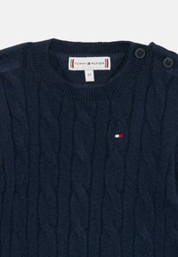 Ναυτικό μπλε πλεκτό πουλόβερ με σχέδιο καλωδίωσης, στρογγυλή λαιμόκοψη και δύο κουμπιά στους ώμους. Διαθέτει μικρό λογότυπο σε κόκκινο, λευκό και μπλε χρώμα.