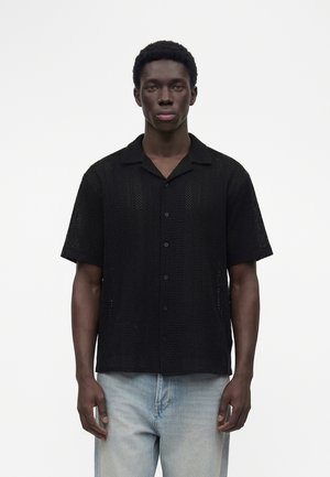 Ung mand med kort krøllet hår iført en sort kortærmet skjorte og lyseblå jeans, stående mod en ensfarvet hvid baggrund.