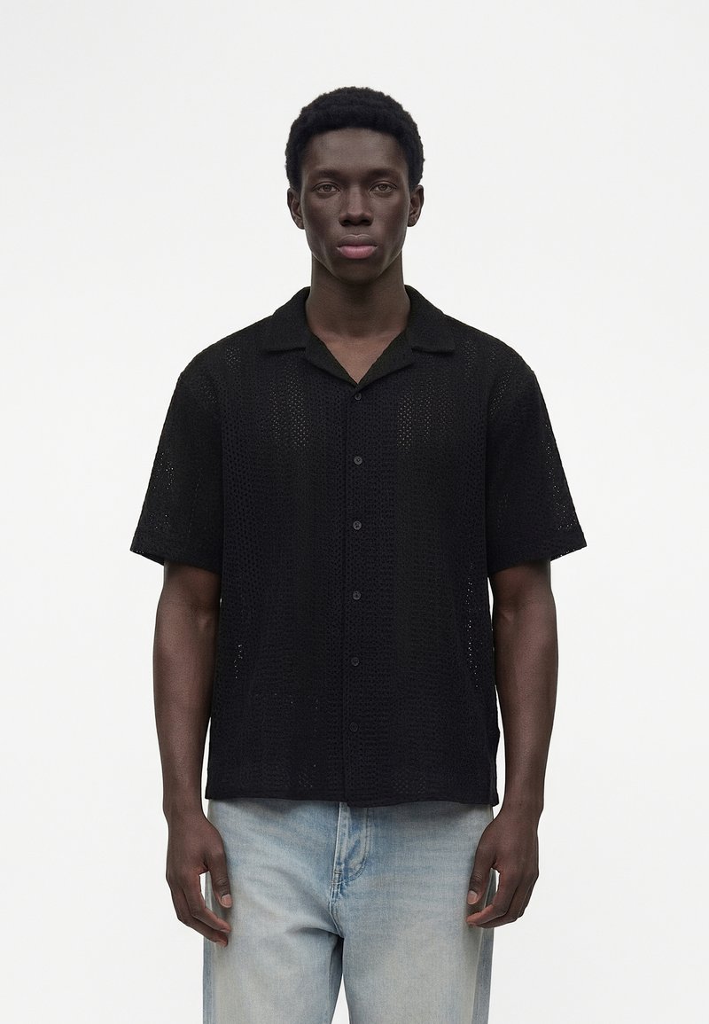 Bărbat tânăr cu păr scurt și creț, purtând o cămașă neagră cu mânecă scurtă cu nasturi și blugi albastru deschis, stând în fața unui fundal alb simplu.