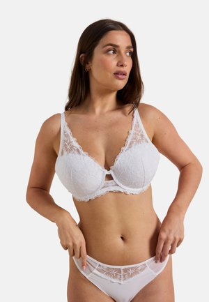 Femme portant un soutien-gorge en dentelle blanche à motifs floraux et une culotte transparente assortie, ajustant sa culotte, regardant sur le côté sur un fond uni.