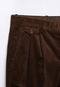 Hnědé kordurové kalhoty s vertikálním ribbed vzorem, zapínáním na knoflík a předními sklady pro zvýšení detailu.