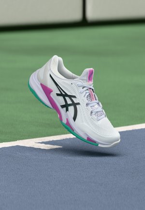 White athletic shoe with black logo, pink and teal accents, suspended above blue and green tennis court surface near the baseline.