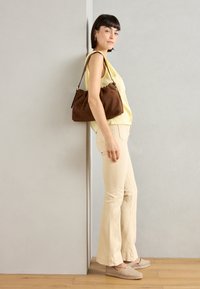 Brun mockaskinnväska med en rynkad design, bärs över axeln. Modell i ljusgul topp, beige byxor och beiga espadriller står mot en vägg.