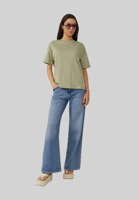 T-shirt oversize vert olive, jeans évasés bleu clair et sandales plateformes crème. Modèle portant de grandes lunettes de soleil, se tenant avec les mains dans les poches.