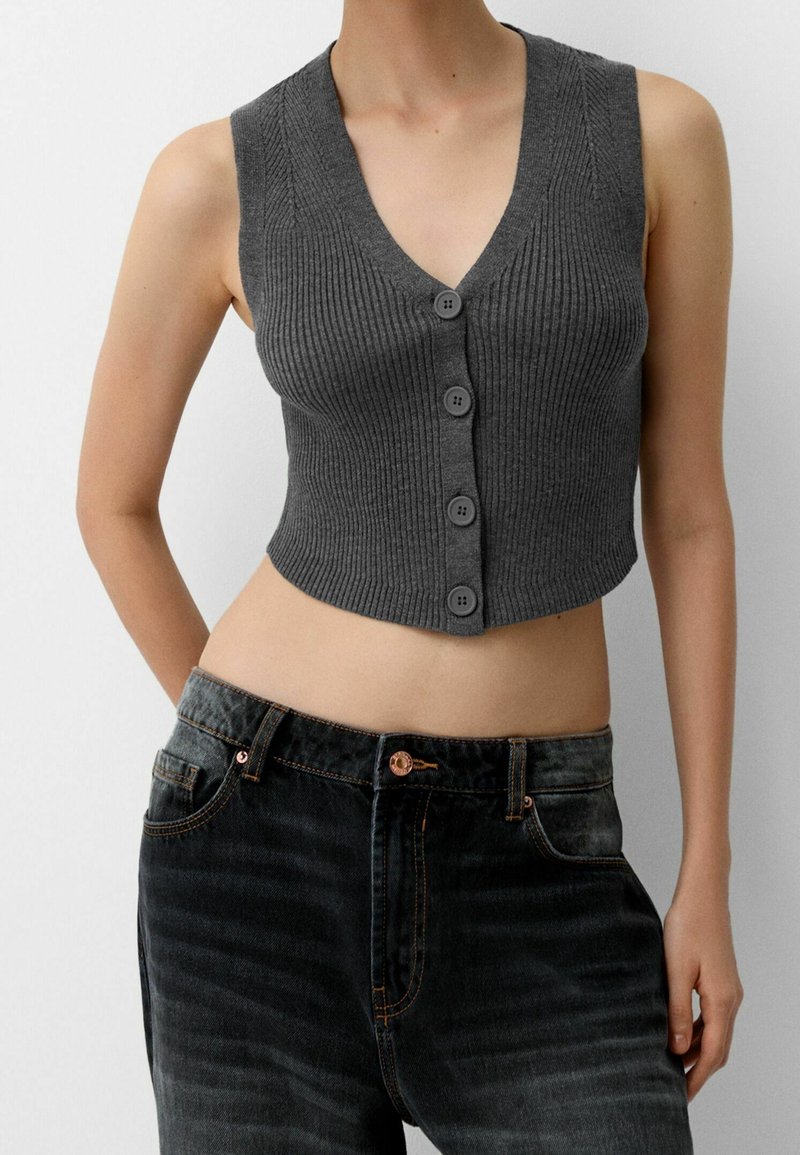 Grå kortärmad stickad topp med djup V-ringning och fem knappslutningar. Bärs tillsammans med högmidjade mörka denimjeans med synlig sömnad.