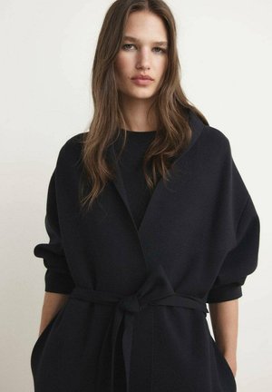 Cappotto avvolgente nero, oversize, realizzato in un tessuto morbido, con cintura da annodare, maniche ampie e una silhouette ampia. Adatto per essere indossato a strati.