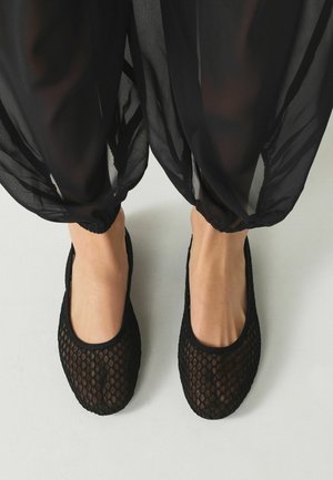 Nohy obuté v čiernych sieťovaných balerínach, skombinované s voľnými, priehľadnými čiernymi nohavicami stiahnutými pri členkoch, stojace na svetlom povrchu.