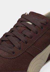 Σκούρο καφέ δερμάτινο αθλητικό παπούτσι από σουέντ με παχιές κορδόνια στο ίδιο χρώμα και μπεζ λεπτομέρεια ραφτών στο πλάι, πάνω σε ανοιχτό φόντο.