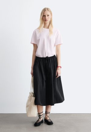 Vrouw draagt een lichtroze T-shirt, zwarte midirok, zwarte veter-ballet flats, houdt een beige boodschappentas vast en staat voor een witte achtergrond.