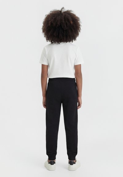 Enfant aux cheveux bouclés, debout de dos, portant une chemise blanche à manches courtes, un pantalon noir et des baskets blanches avec des détails noirs.