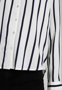 Blouse blanche avec des rayures verticales noires, devant boutonné, manches longues et détail aux poignets associée à un pantalon noir.