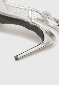 Sølv høyhælte sko med en elegant design laget av glatt metallisk materiale, spiss tå og en tynn, firkantet hæl. Gjennomsiktig stroppdetalj.