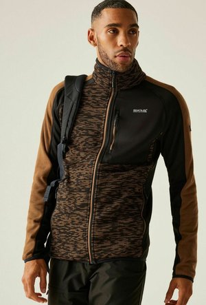 Mann trägt eine braun-schwarze Jacke mit Reißverschluss und Brusttasche und trägt einen schwarzen Rucksackriemen über einer Schulter, schaut nach rechts.