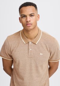 Brown polo shirt made of textured fabric, featuring a collared neck, short sleeves, and contrasting beige trim at the collar and sleeves.