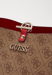 Hnědá texturovaná kabelka se zlatým kováním, výrazným logem "GUESS" a červeným koženým akcentem poblíž stříbrného uzavíracího kroužku.