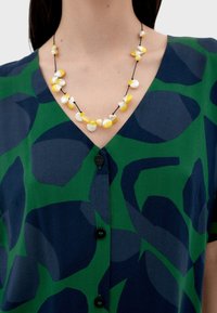 Grün und marineblau gemusterte Bluse mit kurzen Ärmeln und Knopfverschluss, kombiniert mit einer Halskette aus weißen und gelben Perlen an einem schwarzen Band.