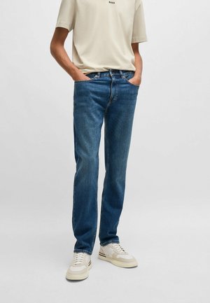 Person iført blå jeans med lige ben, beige T-shirt med "BOSS"-logo og hvide sneakers, står med hænderne i lommerne.