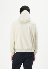 Felpa con cappuccio di un beige chiaro, dal fitting rilassato, con polsini e orlo a costine, e una texture morbida, indossata con pantaloni scuri.