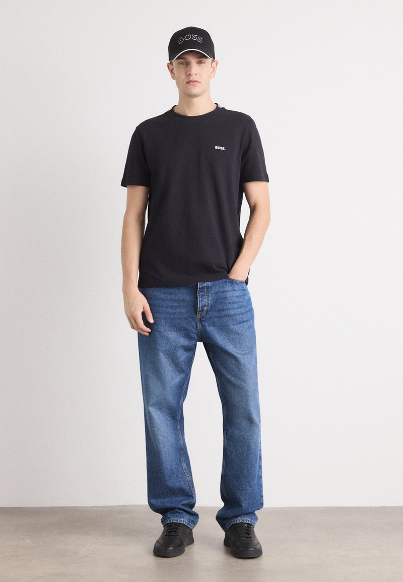 T-shirt en coton noir avec logo blanc, jeans en denim bleu et baskets noires. Le modèle porte une casquette noire avec un logo brodé. Tenue décontractée.