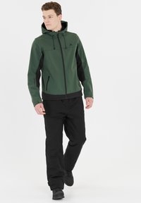 Veste softshell verte avec des accents noirs, poches zippées, capuche ajustable et poignets élastiques. Assortie à un pantalon noir et des chaussures.