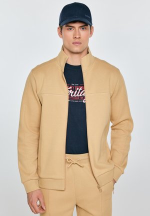 Mężczyzna w beżowej kurtce zapinanej na zamek błyskawiczny i pasujących spodniach, granatowym t-shircie z nadrukiem oraz granatowej czapce baseballowej na jednolitym tle.