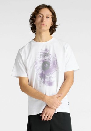 Jonge man met halflang haar, gekleed in een wit T-shirt met een paarse abstracte grafische print en zwarte shorts, staand met gekruiste handen.