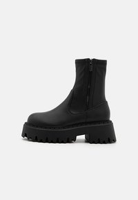 Bottes noires à la cheville en matériau synthétique lisse. Dotées d'une semelle épaisse et crantée ainsi que d'une fermeture éclair sur le côté pour un enfilage facile.