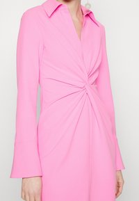 Robe longue rose à manches longues avec un col, présentant un détail avant torsadé et des manches en cloche, fabriquée en tissu lisse avec une finition mate.