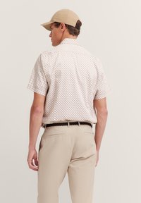 Kurzarmhemd in hellem Beige mit geometrischem Muster, kombiniert mit beigen Hosen und einer sandfarbenen Mütze. Mit einem Standardkragen und geradem Saum.
