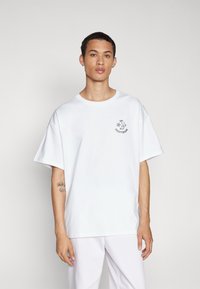 Tânăr îmbrăcat cu un tricou alb oversized, cu un mic logo Converse negru pe pieptul stâng, stând în fața unui fundal simplu, deschis la culoare.