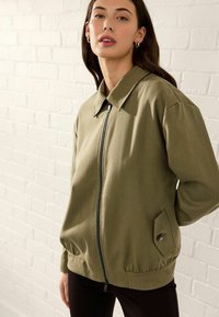 Olivgrüne, Reißverschlussjacke mit Kragen, langen Ärmeln und seitlichen Taschen. Sie verfügt über einen strukturierten Stoff und einen elastischen Bund.