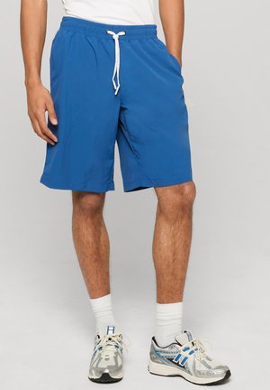 Homme portant un short à cordon bleu, des chaussettes blanches aux chevilles et des chaussures de course argentées et bleues, se tenant debout avec une main dans la poche.