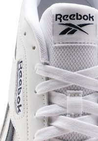Zapatilla blanca de Reebok con parte superior de malla texturizada, detalles de cuero y etiqueta con la marca. Incluye cordones blancos y un logotipo prominente.