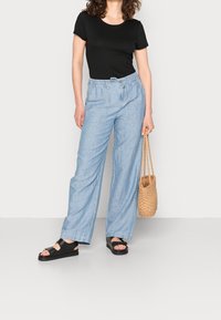 Vrouw in een zwart T-shirt met korte mouwen, lichtblauwe wijde broek, zwarte sandalen, houdend een geweven rieten handtas tegen een effen achtergrond.