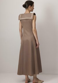 Vrouw met een opgestoken haarstijl in een mouwloze, bruine midi-jurk met een brede vierkante kraag en beige afwerking, gecombineerd met beige slingback schoenen met een lage hak.
