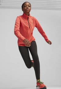 Sportief langemouwen shirt in fel koraal met rits-hals. Gecombineerd met zwarte leggings en rode sportschoenen met groene accenten.