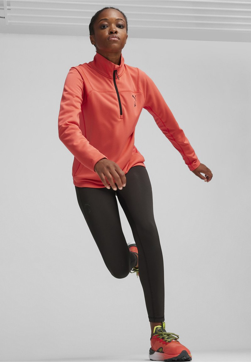 Sportief langemouwen shirt in fel koraal met rits-hals. Gecombineerd met zwarte leggings en rode sportschoenen met groene accenten.