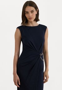 Jūrinės mėlynos suknelės be rankovių, su išsukta priekiu, su sidabriniu grandinėlės akcentu juosmens srityje ir lygia, tekstūrine medžiaga.