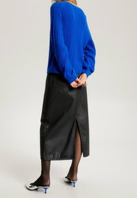 Pull en maille torsadée bleu, jupe mi-longue en cuir noir avec une fente au dos, et mules à talons blanches et bleues. Textures lisses et silhouette ajustée.