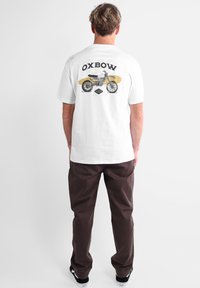 Uomo in piedi di spalle, indossa una maglietta bianca con una grafica di una motocicletta nera e il testo "OXBOW", pantaloni marroni e sneakers nere.