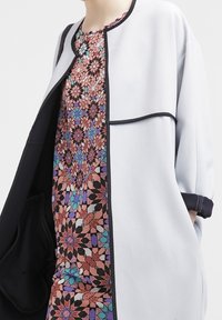 Světlé šedé sako s černým lemováním, nošené přes vzorované šaty s geometrickými motivy v růžových, modrých a fialových odstínech.