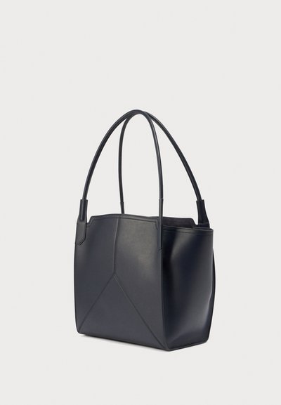 Μαύρη δερμάτινη τσάντα tote με διπλές πάνω λαβές και γεωμετρικό σχέδιο με ραφές στην επιφάνεια, εμφανίζεται διαγώνια σε λευκό φόντο.