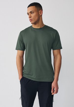 Uomo con camicia verde scuro a maniche corte e pantaloni neri, in piedi con le mani nelle tasche, guarda verso sinistra su uno sfondo semplice.