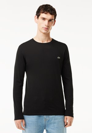 Sort langærmet skjorte lavet af bomuld, med rund halsudskæring og et lille grønt logo på brystet, parret med blå jeans.